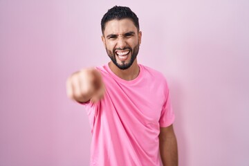 Hispanic young man standing over pink background pointing displeased and frustrated to the camera,...
