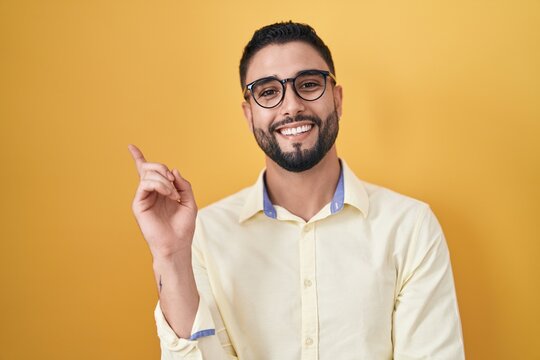 Hispanic young man wearing business clothes and glasses with a big smile on face, pointing with hand finger to the side looking at the camera.