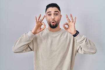 Young handsome man standing over isolated background looking surprised and shocked doing ok approval symbol with fingers. crazy expression