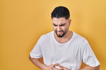 Young handsome man wearing casual t shirt over yellow background with hand on stomach because indigestion, painful illness feeling unwell. ache concept.