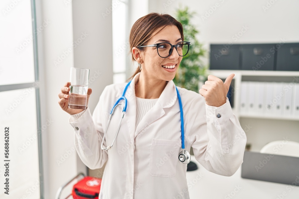 Canvas Prints Young brunette doctor woman holding glass of water pointing thumb up to the side smiling happy with open mouth