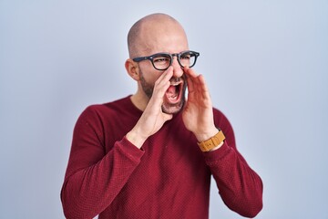 Young bald man with beard standing over white background wearing glasses shouting angry out loud with hands over mouth