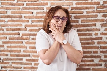 Senior woman with glasses standing over bricks wall touching mouth with hand with painful expression because of toothache or dental illness on teeth. dentist