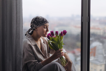 Young woman with cancer sitting in a window and smelling flowers.