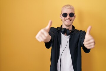 Young caucasian man wearing sunglasses standing over yellow background approving doing positive gesture with hand, thumbs up smiling and happy for success. winner gesture.