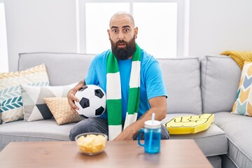 Young hispanic man with beard and tattoos football hooligan holding ball supporting team scared and...