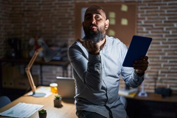 Young hispanic man with beard and tattoos working at the office at night looking at the camera blowing a kiss with hand on air being lovely and sexy. love expression.