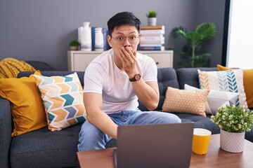 Young asian man using laptop at home sitting on the sofa shocked covering mouth with hands for...