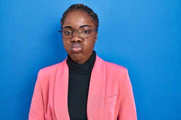 Obraz premium Beautiful black woman standing over blue background puffing cheeks with funny face. mouth inflated with air, crazy expression.