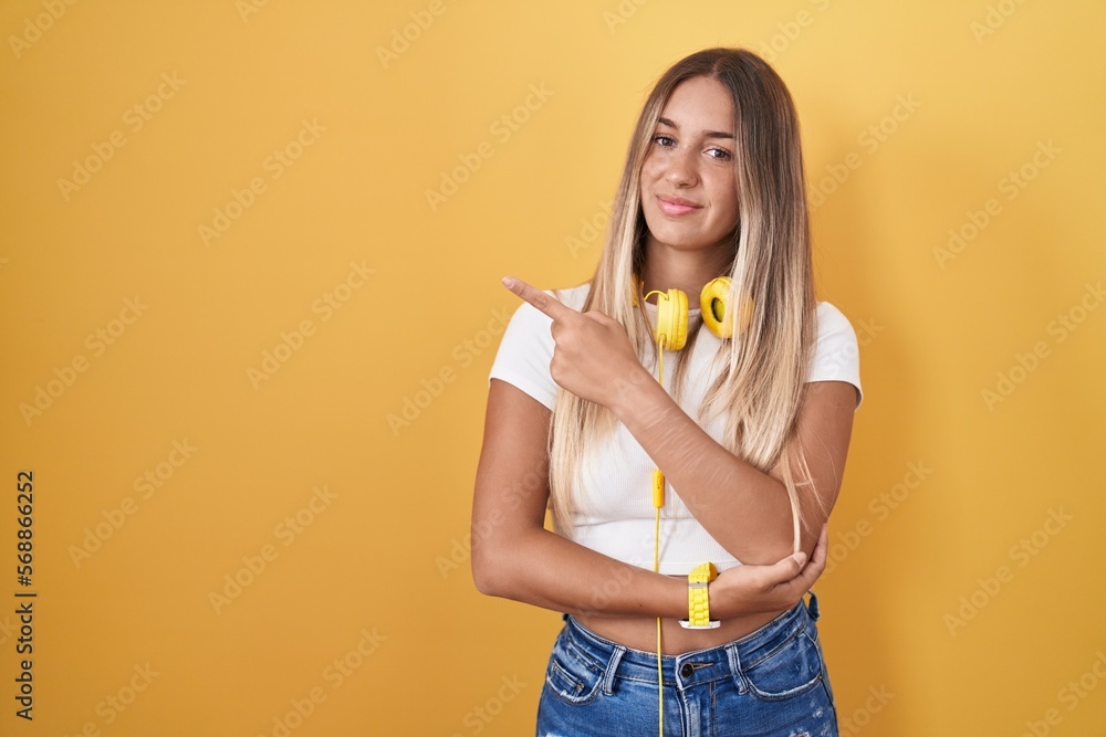 Poster young blonde woman standing over yellow background wearing headphones pointing with hand finger to t