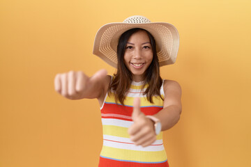 Middle age chinese woman wearing summer hat over yellow background approving doing positive gesture with hand, thumbs up smiling and happy for success. winner gesture.