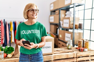 Middle age blonde woman wearing volunteer uniform using smartphone at charity center - Powered by Adobe