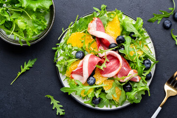 Delicious salad with smoked duck, oranges, blueberries, arugula and lettuce, black table background, top view
