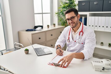 Young hispanic man wearing doctor uniform writing report working at clinic