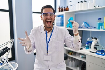 Young hispanic man with beard working at scientist laboratory holding blue ribbon celebrating victory with happy smile and winner expression with raised hands