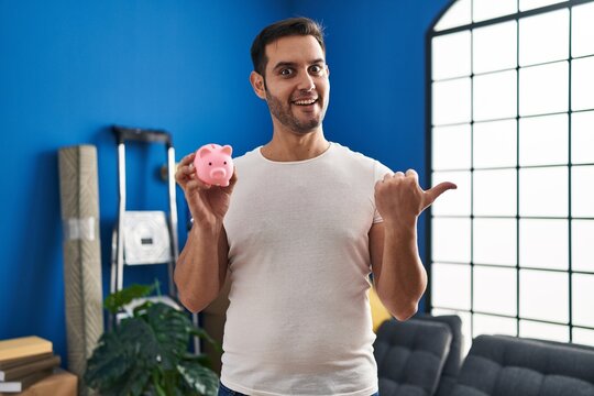 Young hispanic man with beard holding piggy bank at new home pointing thumb up to the side smiling happy with open mouth