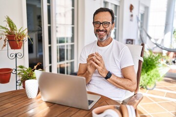 Middle age man using computer laptop at home hands together and fingers crossed smiling relaxed and...