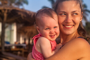 Smiling mom with baby in arms on tropical sandy beach at palm trees background. Female with little child in summer travel vacation. Concept of motherhood and recreation with children. Copy text space
