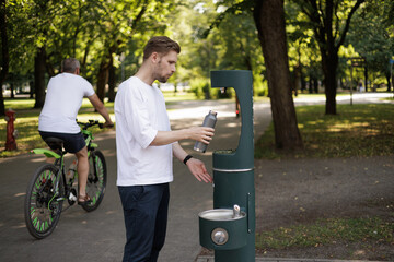 A man in a white shirt refills his water bottle at a public refill station in a sustainable city....