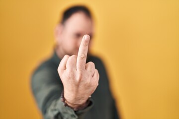 Plus size hispanic man with beard standing over yellow background showing middle finger, impolite...