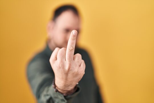 Plus size hispanic man with beard standing over yellow background showing middle finger, impolite and rude fuck off expression