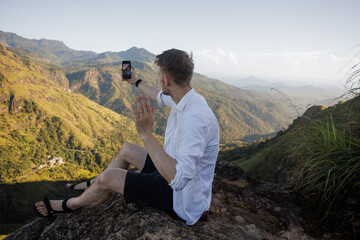 Naklejka premium Young man in nature. People and mountain. Healthy lifestyle. Happy walk in traveling in Sri Lanka. Outdoor activity casual guy with backpack. Male making travel photography with smartphone. Daily life