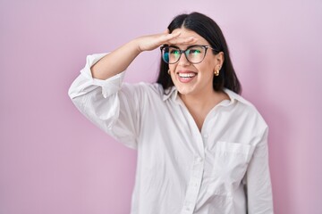 Young brunette woman standing over pink background very happy and smiling looking far away with hand over head. searching concept.