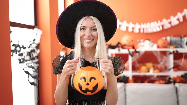 Young blonde woman wearing witch costume holding halloween pumpkin basket at home