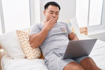 Chinese young man using laptop sitting on the bed covering mouth with hand, shocked and afraid for...