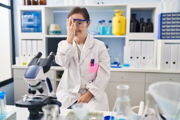 Fototapeta premium Hispanic girl with down syndrome working at scientist laboratory covering one eye with hand, confident smile on face and surprise emotion.