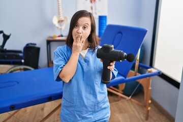 Hispanic girl with down syndrome holding muscle gun at physiotherapy clinic covering mouth with...