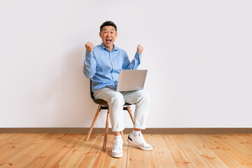 Happy asian businessman sitting on chair with laptop, raising hands up, excited over business success, full length