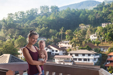 Fototapeta na wymiar Portrait of mom with baby on balkony in tropical country at mountain background. Woman with child in summer travel vacation. Concept of motherhood and family recreation with children. Copy text space