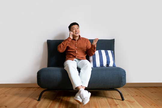 Happy asian middle aged man talking on cellphone, sitting on sofa, having phone conversation against white studio wall