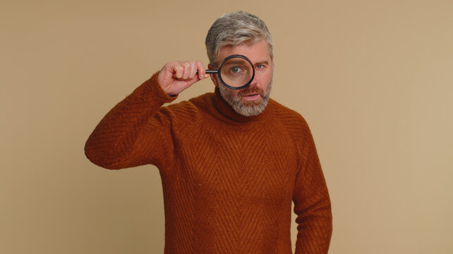 Investigator researcher scientist old man holding magnifying glass near face, looking into camera with big zoomed funny eyes, searching, analysing. Senior middle-aged guy on beige studio background