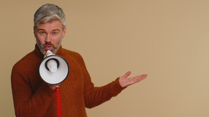 Middle-aged man talking with megaphone, proclaiming news, loudly announcing advertisement, warning using loudspeaker to shout speech, pointing empty place. Copy-space. Senior guy on beige background