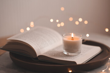 Burning scented candle staying on open paper book on wooden tray over glow lights on table in bedroom. Cozy home atmosphere.