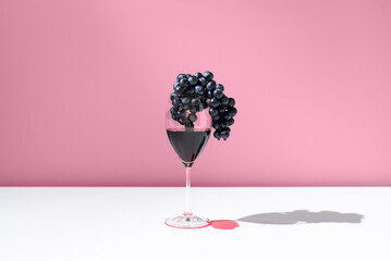 Un racimo de uvas negras dentro de una copa de vino tinto goteando sobre fondo rosa