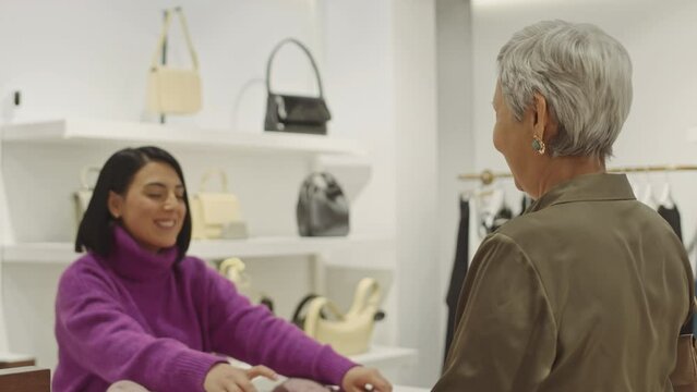 Elegant mature Asian woman shopping for clothes in luxury designer showroom, giving purchased items to female shop assistant at counter