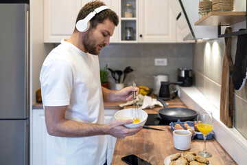 Fototapeta na wymiar Man in white tee shirt and headphones standing in kitchen cooking omelet for breakfast and listening music.