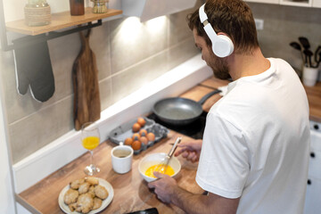 Man in white tee shirt and headphones standing in kitchen cooking omelet for breakfast and...