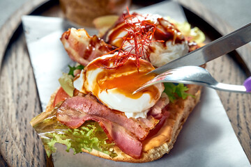 A girl cuts a sandwich with tomato, bacon and a poached egg for breakfast. Concrete background. Hard light