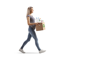Full length profile shot of a young woman carrying a cardboard box with recycling plastic bottles and walking