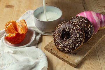  Donuts with chocolate and pink frosting over a wooden table, milk cup and spilled milk, white towel and a mandarin