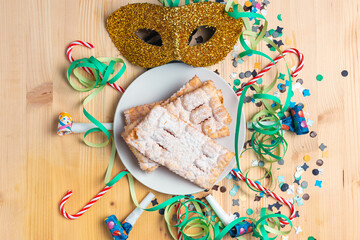 Top view of a plate of Chiacchiere over a wooden table with carnival trumpet, carnival mask, confetti, streamers and candy canes