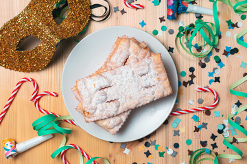 Top view of a plate of Chiacchiere over a wooden table, with carnival trumpet, carnival mask, confetti, streamers and candy cane