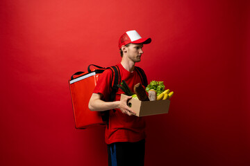 Side view delivery man in red uniform on a red background delivering food, groceries, vegetables, drinks in a paper box to a client at home. Online grocery shopping service concept.