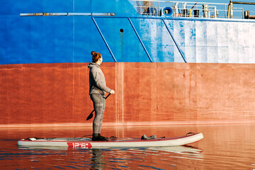 Fototapeta na wymiar Woman on stand up paddle board (SUP) rowing on the river near cargo ship at autumn