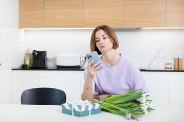 Young attractive woman with bouquet of flowers and gift box sitting in the kitchen and looking at her smartphone