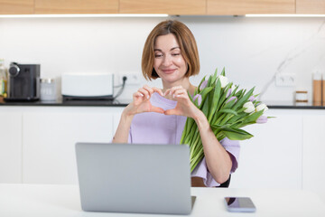 Young happy woman with bouquet of flowers sitting at the table and talking on video call on laptop. Online dating and  celebrating holidays far from each other concept